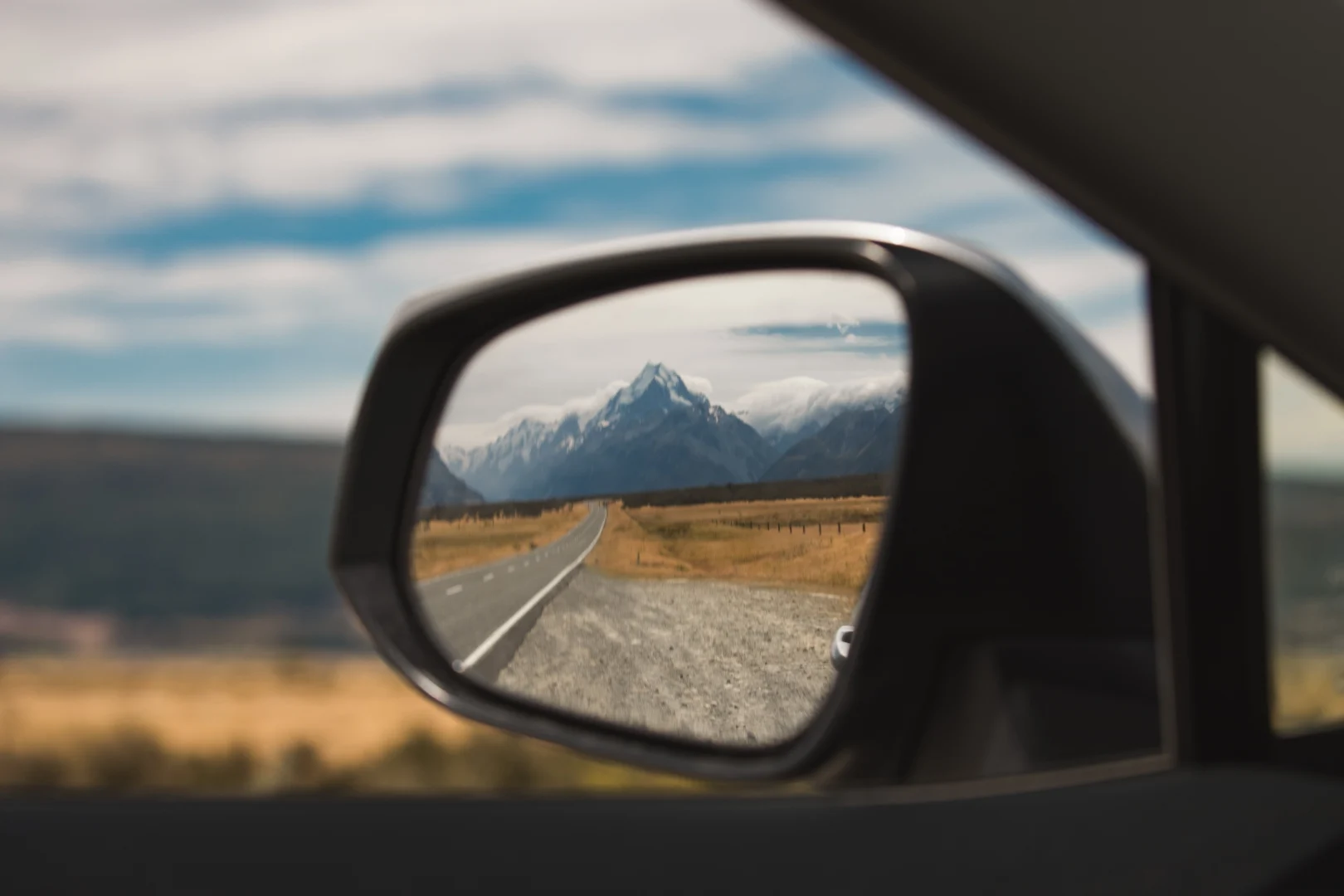 Reflection of snow-capped mountains and a road in a car's side mirror